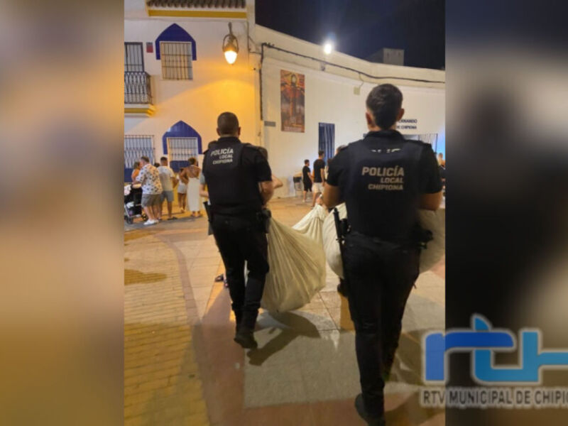 Dos policías locales de Chipiona, Andalucía, llevan una bolsa mientras se observa a un grupo de personas en la calle. La imagen parece ser tomada durante una noche, con luces y un edificio de fondo.