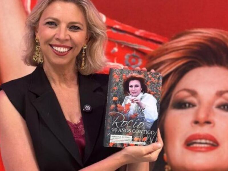 Una mujer sonríe mientras sostiene un libro titulado 'Rocio: 20 años contigo' frente a una imagen de la actriz Rocio Durcal.
