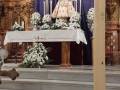Una imagen de una iglesia con un altar adornado con flores y velas encendidas. En el centro, una estatua de la Virgen María rodeada por flores y velas. En el fondo, detalles arquitectónicos elaborados.