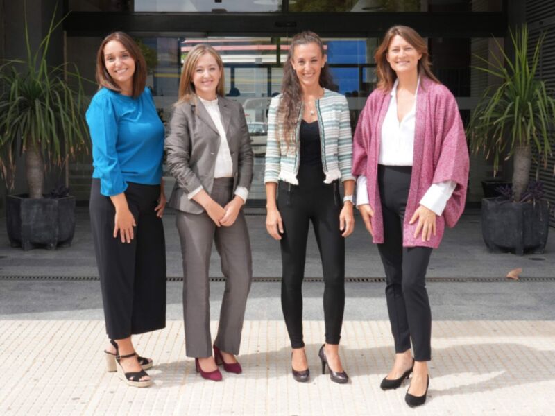 Cuatro mujeres en trajes de negocios posan frente a un edificio moderno. La imagen muestra una escena formal y profesional, probablemente en un entorno de oficina o evento corporativo.