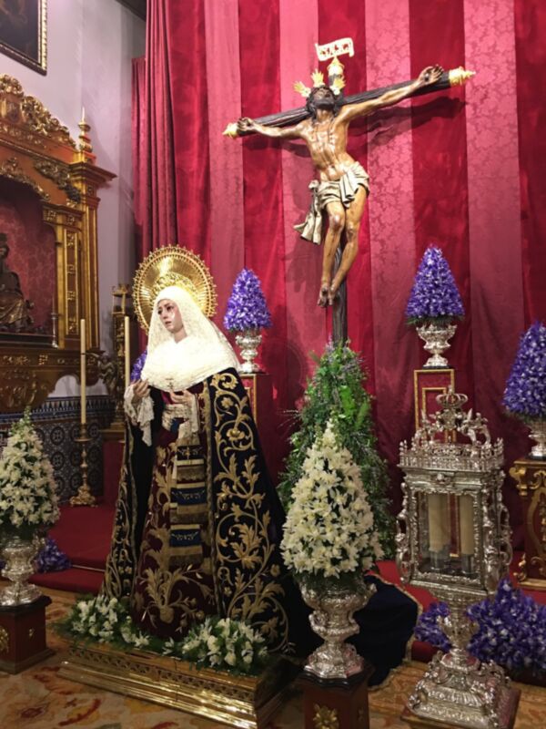 Imagen religiosa con una Virgen María vestida en traje negro y dorado, posando con un niño en sus brazos. En el fondo, una cruz con Cristo crucificado y cortinas rojas. Arriba de la cruz, un escudo heráldico dorado y plateado. En el suelo, adornos florales y una alfombra de color rojo con detalles dorados.