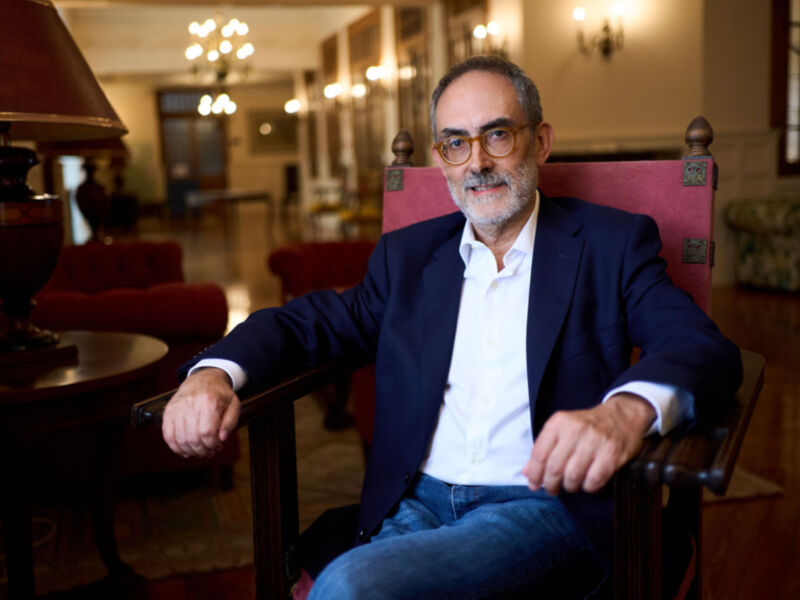 Un hombre con una camisa blanca y pantalones vaqueros se sienta en un sillón rojo, con una lámpara de mesa a su lado. La habitación tiene un estilo clásico con cortinas y una chimenea en el fondo.