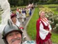 Un grupo de personas vestidas con trajes históricos y tocados se desplaza por un camino en un parque. La escena está rodeada de flores y árboles, con un cielo nublado.