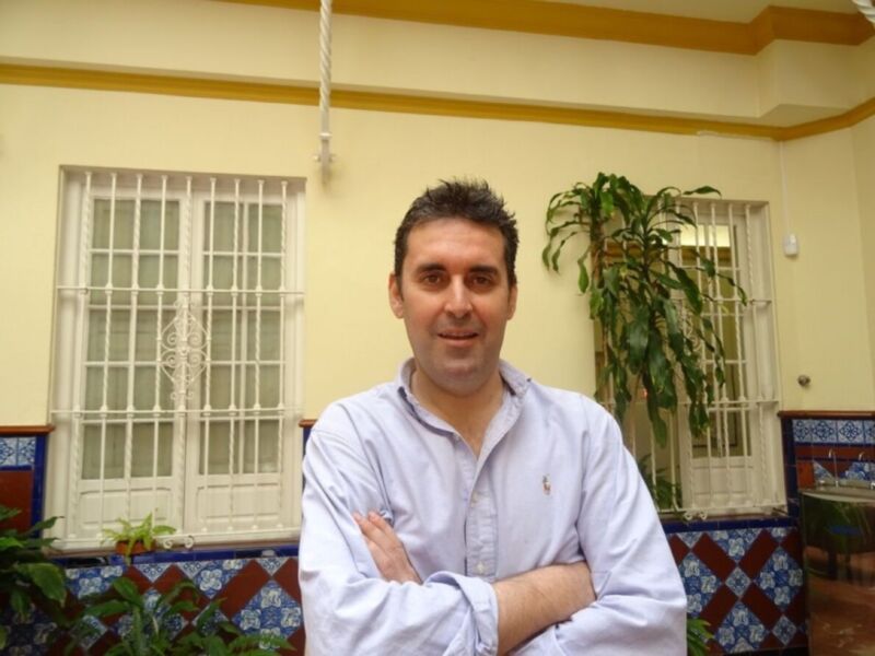 Un hombre con camisa blanca y cabello corto, posando en una habitación con paredes amarillas y ventanas de barrotes. Hay un rincón decorado con azulejos coloridos y una planta en la esquina.