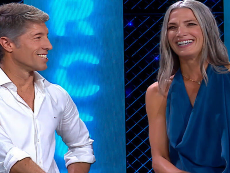 Dos personas sonríen en un escenario con fondo azul y luces. La persona de la izquierda lleva una camisa blanca, mientras que la de la derecha usa un vestido azul.