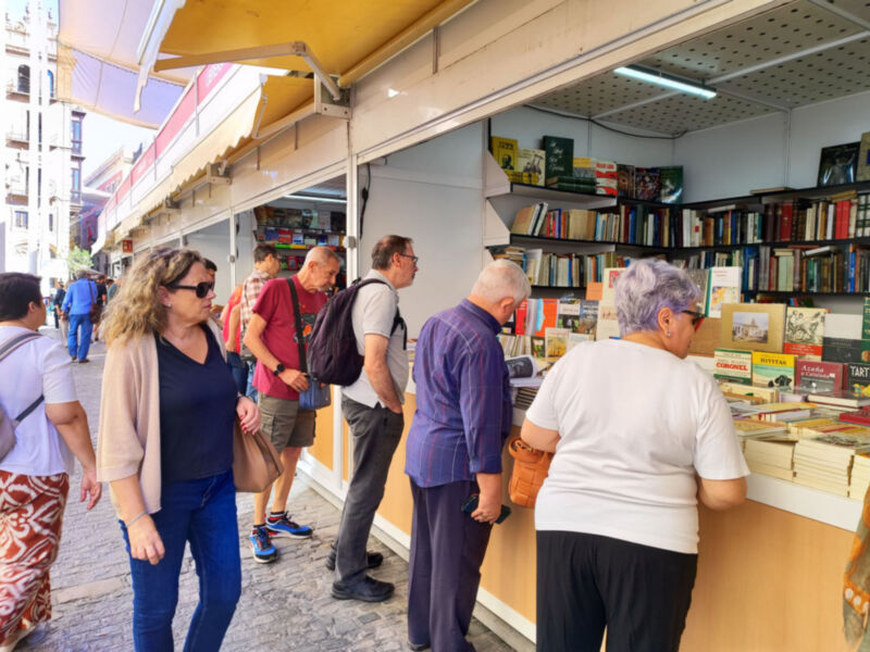 Una librería al aire libre con clientes esperando en línea. La imagen muestra a varias personas, algunos de ellos leyendo libros o conversando entre sí. El exterior está iluminado con luz natural y el interior parece ser un espacio amplio con estanterías llenas de libros. La atmósfera es tranquila y relajada, típica de un día soleado en la ciudad.