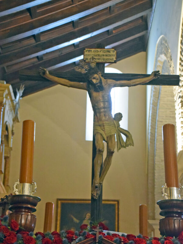 Crucifijo en una iglesia con detalles de madera y velas encendidas. Arriba, un cuadro con una figura en un fondo oscuro. Abajo, flores rojas y ramas de olivo. Contexto religioso y decorativo.
