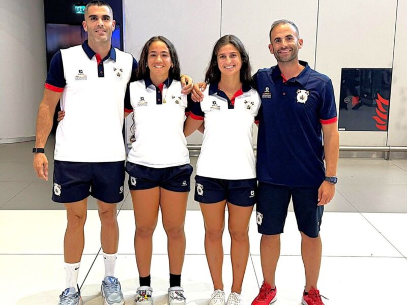 Tres personas posan juntas, dos de ellas son mujeres y una es hombre. Todos llevan uniformes deportivos con logos distintos, probablemente representando un equipo o organización. El fondo es interior y moderno con elementos de diseño minimalista.