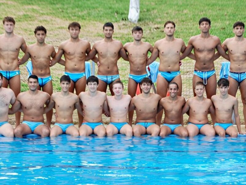 Un grupo de hombres en trajes de baño posando frente a una piscina azul. Algunos llevan camisetas negras y otros tienen pantalones cortos azules. La escena está en un área verde con césped y una pared de madera.