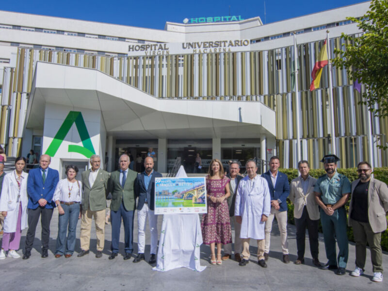 Se muestra un grupo de personas frente a una entrada principal de un hospital universitario, con una bandera y un cartel en el centro. El cielo es azul claro y hay árboles al lado del edificio.