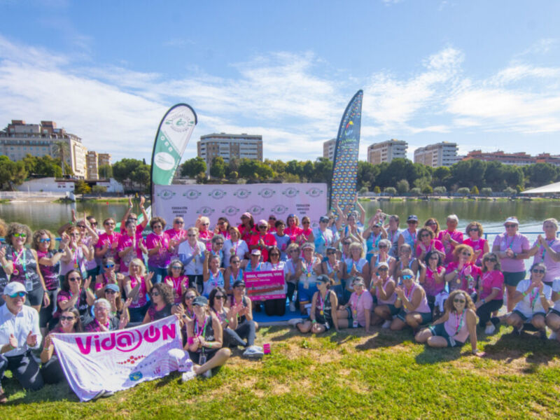 Una gran cantidad de personas con camisetas rosas se reúnen en un parque junto a un lago. Algunos están sentados, otros se inclinan para tocar el suelo o posar. En la parte superior de la imagen, hay dos banderas con la palabra "VIDOJUN" en letras grandes. El cielo es azul claro y el paisaje incluye edificios, árboles y un par de toldos.