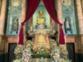Una estatua de la Virgen María vestida en traje dorado, rodeada por flores y velos rojos. La estatua está colocada en un altar con columnas de mármol y una arquitrabe decorado. El entorno es de estilo barroco, con detalles ornamentales y un cielo azul claro.