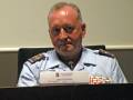El hombre está sentado en una silla de conferencia, con un nombre cartel en frente. Lleva una chaqueta militar y un insignia de rango en el pecho izquierdo. La imagen parece ser de una conferencia o reunión formal.