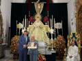 En una iglesia, un hombre y una mujer sostienen fotos mientras un sacerdote se sienta en un trono. La imagen está rodeada de flores y velas, con una estatua central que parece la Virgen María.