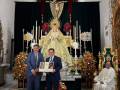 Dos hombres en trajes de negocios sostienen un libro mientras una persona vestida con un hábito religioso se sienta en la derecha. La imagen muestra una escena formal con elementos religiosos y decorativos, incluyendo un altar con una imagen de la Virgen María y el Santo Cristo. La decoración incluye flores y una alfombra con patrones geométricos en el suelo.