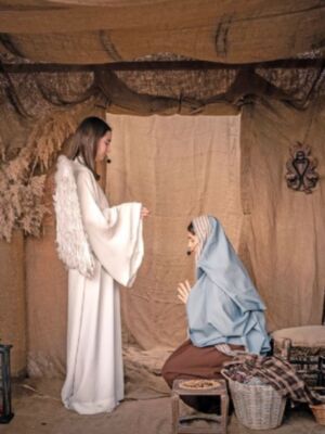 En una escena navideña, dos personajes vestidos con trajes tradicionales se encuentran en un entorno de madera y tela. La figura de pie, con una capa blanca y alas, posiblemente representa a un ángel, mientras que la otra, con una saya azul y tocado, podría interpretar a María. La escena transmite un ambiente festivo y religioso.