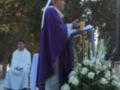 El sacerdote, vestido con un manto púrpura y una mitra blanca, se dirige a la audiencia desde un podio. A su lado, hay una mesa con flores blancas y un microfono. La escena se desarrolla en un entorno al aire libre con árboles y una persona vestida de blanco sentada en el fondo.