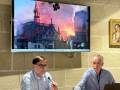 Dos hombres en una sala de conferencias, uno frente a un televisor que muestra imágenes de Notre-Dame en llamas.