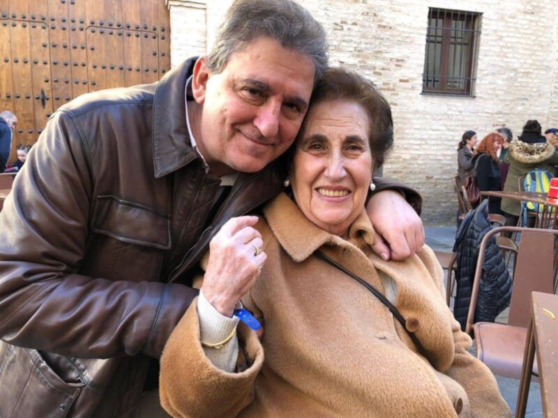 A una pareja abrazándose en un ambiente exterior. La mujer está sonriendo y la mujer tiene una sonrisa amable. Ambos están vestidos con abrigos de invierno. En el fondo, se pueden ver sillas y una mesa con algunos objetos en ella. La pared de la casa tiene un portón de madera y una ventana con persianas negras.